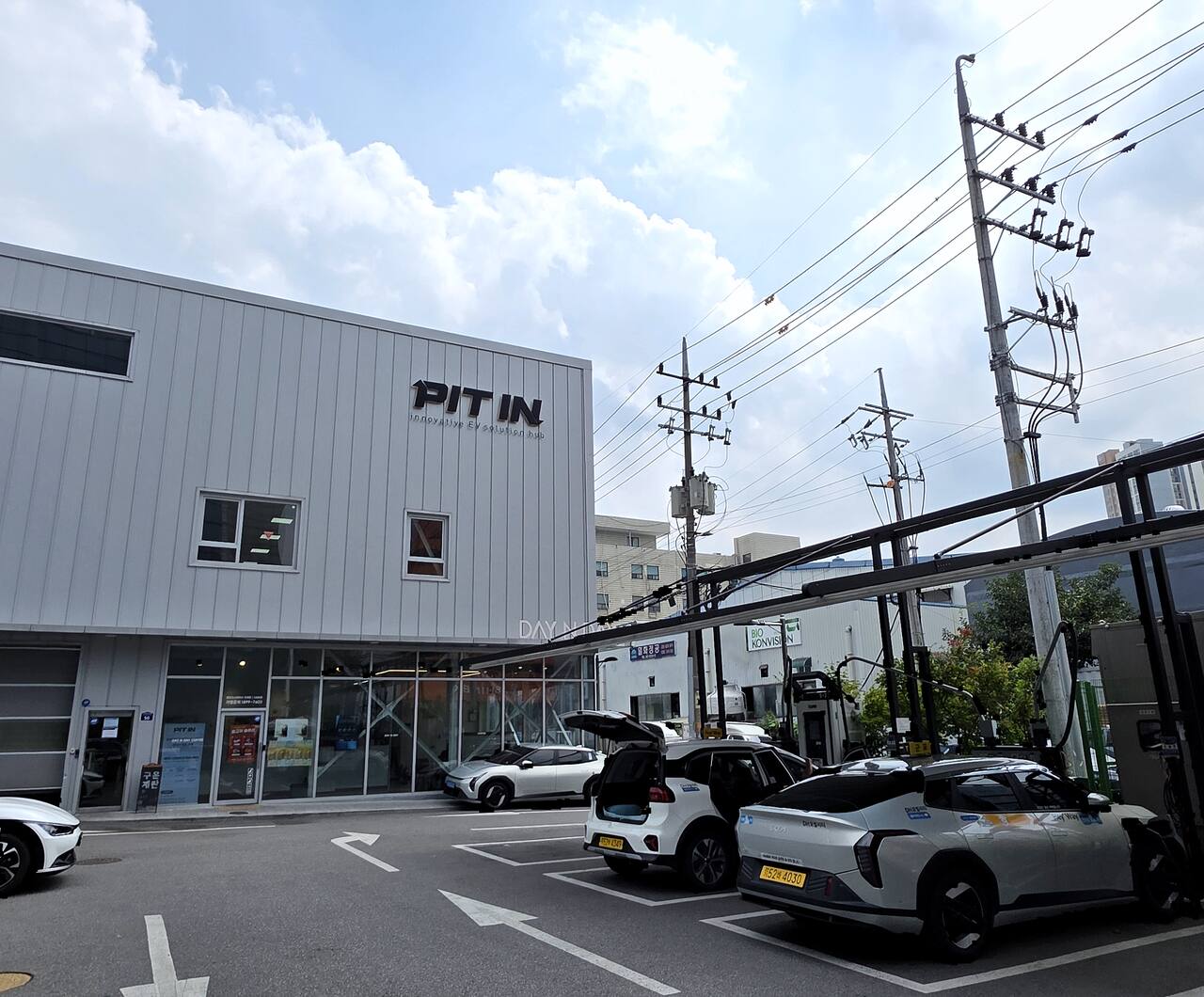 피트인 안양 스테이션 전경 — PIT IN 입체 사인을 얹은 정비동과 유리 로비, 오른쪽 충전 캐노피 아래 충전 중인 전기차들